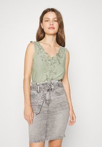 Top sem mangas verde claro com bordados florais no decote, combinado com uma saia de ganga cinza de cintura alta com um cinto atado.