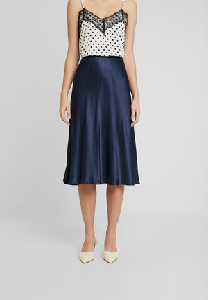 Frau trägt einen marineblauen Satin-Midirock, ein weißes, gepunktetes Top mit schwarzer Spitzenborte und cremefarbene spitze Absätze vor einfachem grauem Hintergrund.