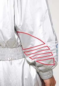 Chaqueta bomber plateada con puños y dobladillo elásticos, adornada con acentos bordados en rojo y azul, con una textura lisa y brillante.