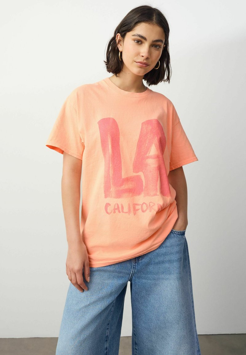Lys orange bomulds t-shirt med fed pink "LA" tekst og "CALIFORNIA" i et afslappet skrifttype. Korte ærmer og løs pasform. Sættes sammen med vide jeans.