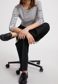 Maglietta a maniche lunghe a righe bianca e nera, abbinata a pantaloni larghi neri e mocassini neri, seduta su una sedia da ufficio.