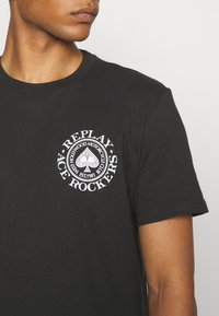 Czarny t-shirt bawełniany z okrągłym białym logo graficznym z tekstem oraz wzorem pika na lewej stronie klatki piersiowej.