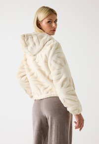 Giacca con cappuccio in pelliccia sintetica color crema con motivi testurizzati a onde; silhouette aderente; abbinata a pantaloni in morbido tessuto grigio.