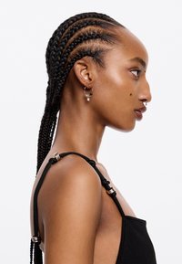 Top senza maniche nero con spalline annodate e accenti metallici. Vista di profilo che mostra un'acconciatura intrecciata e orecchini a cerchio con charm decorativi.