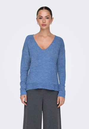 Femme portant un pull bleu à col en V et un pantalon large gris foncé, debout face à l’avant sur un fond uni.