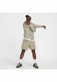 Sudadera con cremallera de color gris claro y tela texturizada, combinada con pantalones cortos beige y zapatillas deportivas grises. El modelo está estirando un brazo hacia afuera.