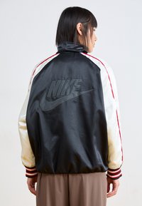 Crna satenska bomber jakna s bijelim rukavima, crvenim prugama i utisnutim Nike logom na leđima. Rebrasti manžeti i pojas.