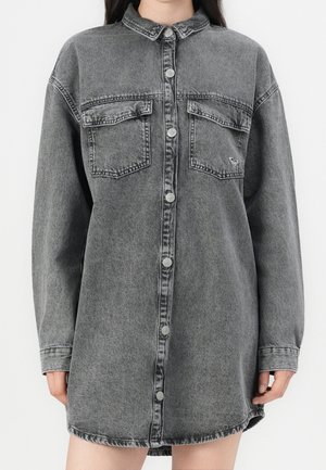 Długa sukienka-koszula z czarnego, wyblakłego denimu z długim rękawem, zapinana z przodu na zatrzaski, z dwoma kieszeniami na klatce piersiowej z patkami, noszona przez osobę.