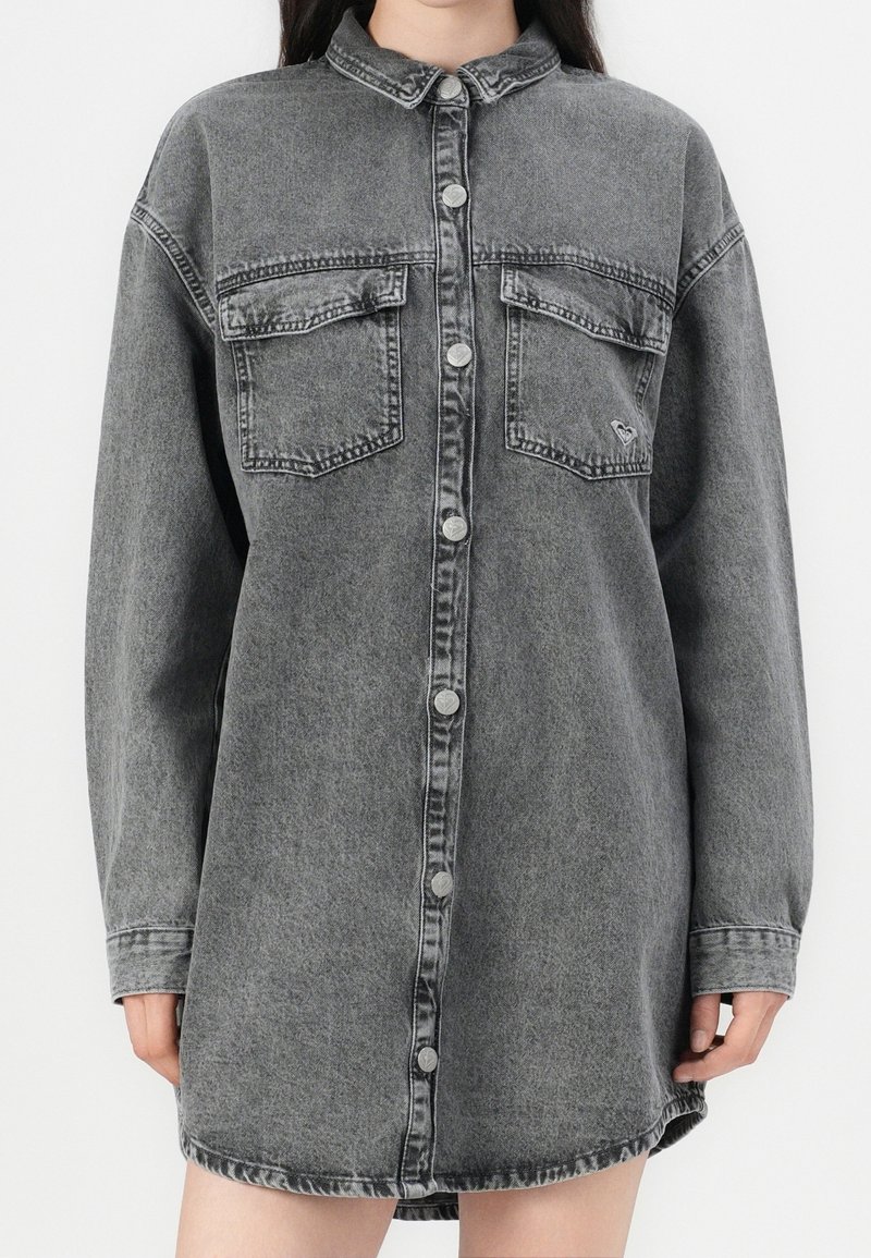 Robe chemise en denim noir délavé à manches longues, avec boutons-pression à l'avant et deux poches à rabat sur la poitrine, portée par une personne.