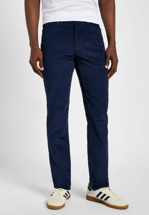 Pantaloni slim-fit blu scuro con una texture liscia, design a cinque tasche e dettagli di cucitura discreti, abbinati a sneaker bianche con strisce nere.