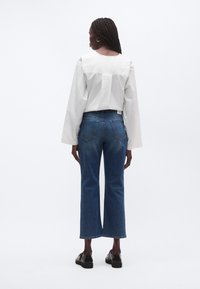 Fehér, rövidített szabású blúz hosszú ujjakkal, közép-kék farmerrel párosítva, mely egyenes szabású, jellegzetes hátsó zsebrátétekkel.