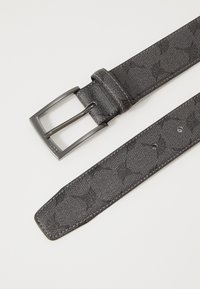 Černý pás zdobený vzorem, vyrobený z texturovaného materiálu, s obdélníkovou kovovou přezkou a několika otvory pro nastavení podél popruhu.