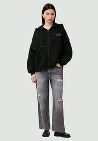 Juodas zip-up megztinis su gobtuvu ir logotipu ant krūtinės, derinamas su nusidėvėjusiais, plačiais pilkais džinsais ir juodais auliniais batais.