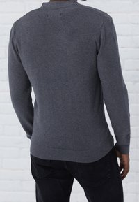 Mann in einem dunkelgrauen Strickpullover und schwarzen Jeans, der mit dem Rücken zur Kamera vor einer weißen Ziegelwand steht.