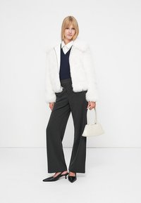 Giacca in pelliccia sintetica bianca, maglione blu navy, pantaloni a righe nere, decolletè neri a punta e una piccola borsa bianca. Sfondo semplice.