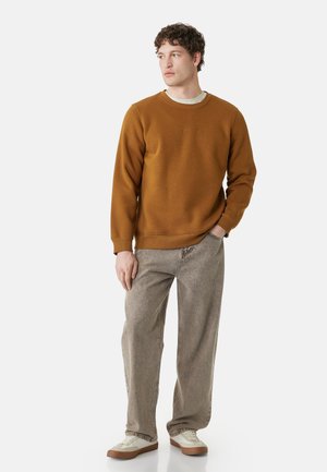 Mann mit braunem Sweatshirt, hellgrauen Jeans und weißen Sneakers, steht mit einer Hand in der Tasche und schaut zur Seite.