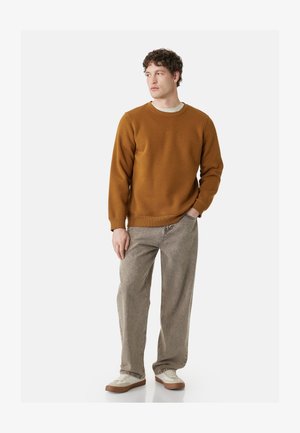 Uomo con felpa marrone, jeans grigio chiaro e scarpe da ginnastica bianche, in piedi con una mano in tasca, guarda di lato.