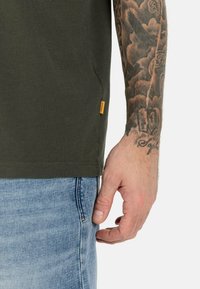 Olivengrøn kortærmet t-shirt med blød tekstur, der har et lille orange mærke på siden, parret med lyseblå denim.