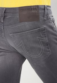 Šedé denimové džíny s logem True Religion na nášivce, s charakteristickým prošíváním zadních kapes do tvaru U a slim fit designem.