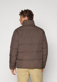 Brun pufferjakke med høj krave, quiltet design og elastiske manchetter. Glat tekstur, let lysende finish, båret over beige bukser.
