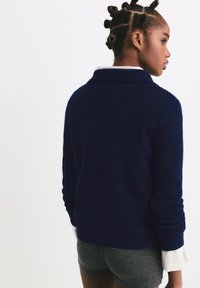 Marineblauer Strickpullover mit Schalkragen, gerippten Bündchen und strukturiertem Finish, kombiniert mit grauen Shorts und einem gestreiften weißen Hemd.