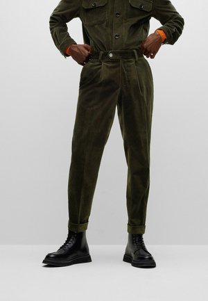 Person trägt hoch taillierte olivgrüne Cordhosen mit Umschlägen, passende Jacke und schwarze Schnürstiefel, passt die Hose in der Taille an.
