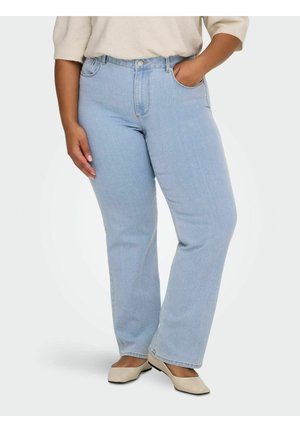 Personne portant un jean droit bleu clair, un pull beige et des chaussures plates beiges, debout avec une main dans la poche.