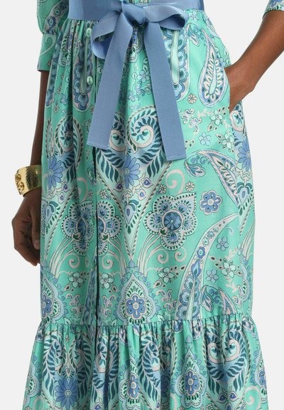 Hand wearing gold bracelet next to turquoise dress with blue floral paisley pattern and tied blue fabric belt at waist.