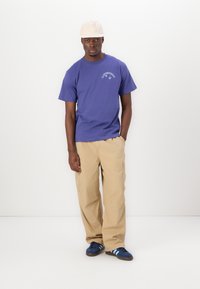 Lilla bomull T-skjorte med en logo nær halsen, sammen med beige cargo bukser. Personen har på seg en lysfarget caps og blå sneakers.