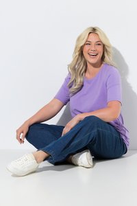 Femme blonde en chemise violette et jeans assise en tailleur sur le sol, souriante et regardant sur le côté.