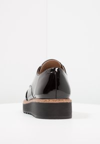 Chaussure en cuir verni noir avec un bout rond, accent en liège sur la semelle et semelle extérieure en caoutchouc noir texturé. Fini lisse avec des coutures minimales.