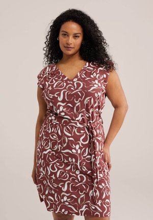 Göndör hajú nő, aki ujjatlan, övvel ellátott rozsda színű ruhát visel fehér absztrakt virágmintával, semleges háttér előtt áll.