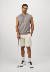  bezrękawnik w kolorze szarym z dekoltem w serek, kremowe szorty, białe buty golfowe z granatowymi akcentami, trzymający kij golfowy, pozujący na jednolitym tle.