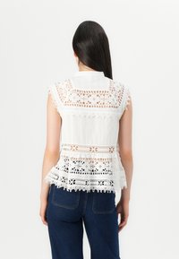 Top bianco senza maniche con dettagli in pizzo intricati, colletto e orlo peplum. Il tessuto è leggero e testurizzato.