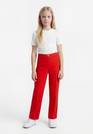 Chica joven con pelo largo y rubio lleva una camiseta blanca de manga corta, pantalones rojos de talle alto con una hebilla dorada y zapatillas blancas, de pie.