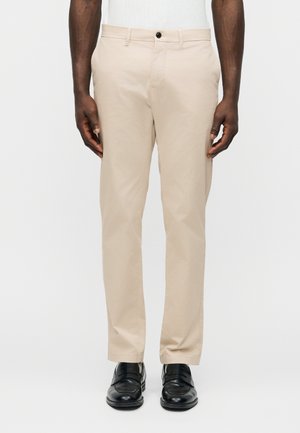 Mand iført beige bukser med lige ben, sorte finsko og en hvid skjorte foran en ensfarvet hvid baggrund.