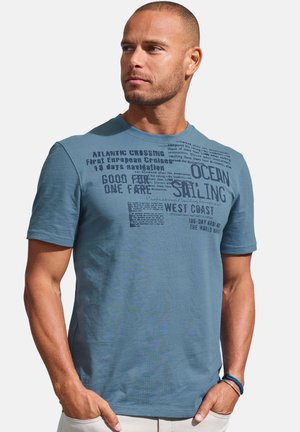 Man with short hair and beard wearing blue sailing-themed t-shirt and light pants, standing with hands in pockets, looking right.