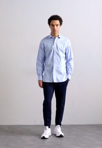 Světle modrá košile na knoflíky, tmavě modré kalhoty a bílé tenisky. Košile má límec a dlouhé rukávy; kalhoty jsou slim-fit.