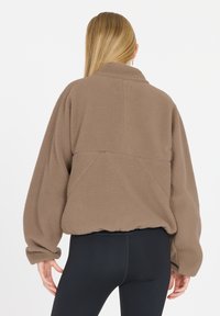 Braune Fleecejacke mit hohem Kragen, fallenden Schultern und elastischen Bündchen. Getragen mit schwarzen Leggings, die eine lässige Passform und Textur zeigen.