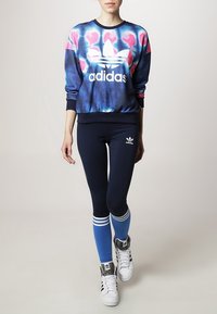 Mikina Adidas s modro-růžovým tie-dye vzorem, spárovaná s tmavě modrými legínami se modrými pruhy a bílými akcenty, a bílými teniskami.