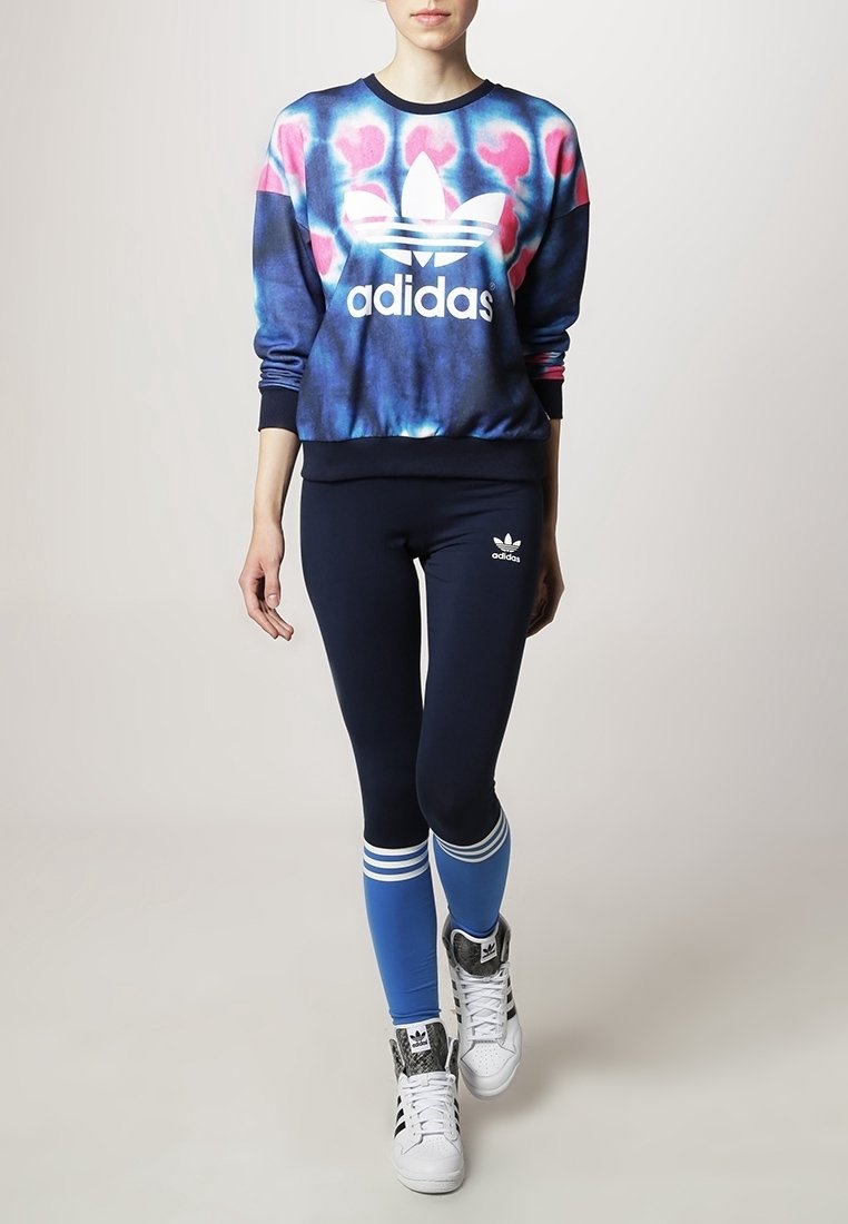 Mikina Adidas s modro-růžovým tie-dye vzorem, spárovaná s tmavě modrými legínami se modrými pruhy a bílými akcenty, a bílými teniskami.