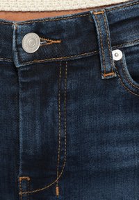 Tmavomodré rifle z denimu so strieborným gombíkom a kontrastným prešitím. Detailný záber pásu ukazujúci hladkú textúru a detaily vrecka.