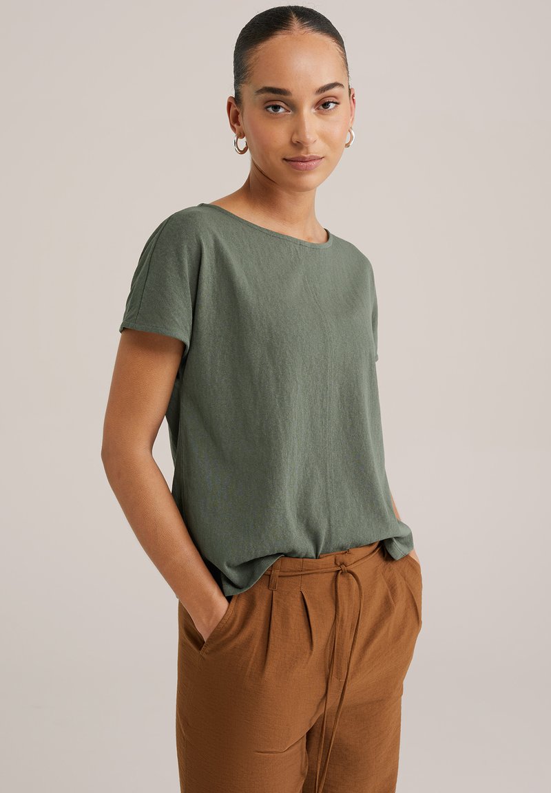 Īsām piedurknēm olīvu zaļš T-krekls, izgatavots no mīksta, elpojoša auduma. Tam ir apaļa kakla izgriezums un brīvs piegriezums. Apvienots ar brūnām teksturētām biksēm.