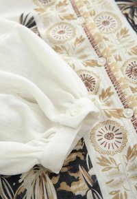 Blouse à manches longues en tissu crème avec des motifs floraux beiges et bruns. Dotée d'une fermeture à boutons et de détails de manches froncées.