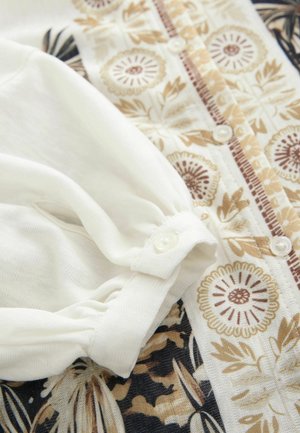 Blouse à manches longues en tissu crème avec des motifs floraux beiges et bruns. Dotée d'une fermeture à boutons et de détails de manches froncées.