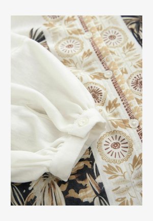 Blusa a maniche lunghe in tessuto crema con motivi floreali in beige e marrone. Presenta una chiusura con bottoni e dettagli di maniche arricciate.