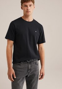 Jaunas vyras vilkintis paprastą juodą trumpomis rankovėmis marškinėlį su mažuoju baltu logotipu, pilkais džinsais ir juodu diržu, stovi priešais kreminį foną.