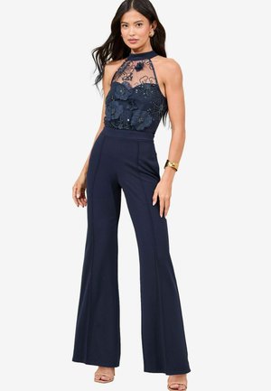 Marineblauer Jumpsuit mit einem transparenten, verzierten Oberteil mit floralem Applikationen, hohem Kragen und weitem Bein, aus einem glatten Stoff gefertigt.