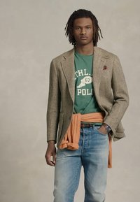 Jovem com dreads, vestindo um blazer de tweed castanho, uma t-shirt gráfica verde, um suéter laranja amarrado na cintura e jeans azul claros.