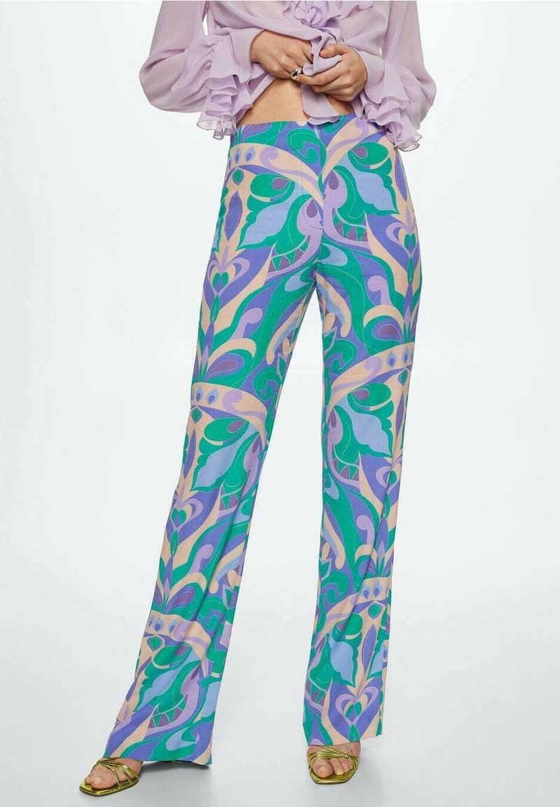 Femme portant un pantalon taille haute avec un motif abstrait coloré de feuilles en vert, violet et beige, assorti à des sandales dorées ouvertes à l’orteil.